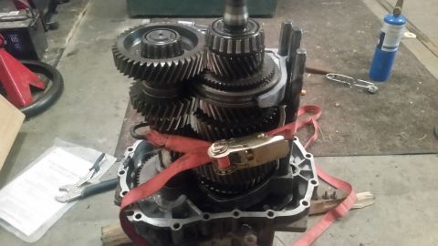 1985 Ford Bronco - A close-up view of a manual transmission's internal gears and components, secured with a red ratchet strap. Tools and other garage items ar…