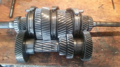 1985 Ford Bronco - A disassembled manual transmission lays on a wooden workbench. The transmission contains multiple gears and shafts, with helical cut teeth…