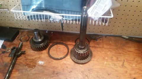 1985 Ford Bronco - Several transmission gears and shafts are laid out on a wooden workbench. A wire rack with some packaging is visible in the background.