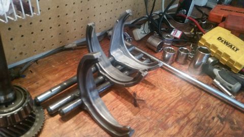 1985 Ford Bronco - Several metal shift forks and other automotive parts are laid out on a wooden workbench next to various tools, including socket wrenches an…