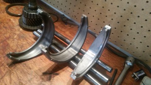1985 Ford Bronco - Several metal bearing races are laid out on a wooden workbench, resting on metal rods. In the background, a transmission gear set is visibl…