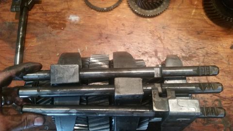 1985 Ford Bronco - A mechanic's hands are shown holding a transmission input shaft with gears attached. The shaft is resting on a wooden surface with other tr…