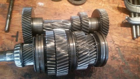 1985 Ford Bronco - Two transmission input and output shafts with multiple gears are laid out on a wooden surface. The shafts are made of metal and have helica…