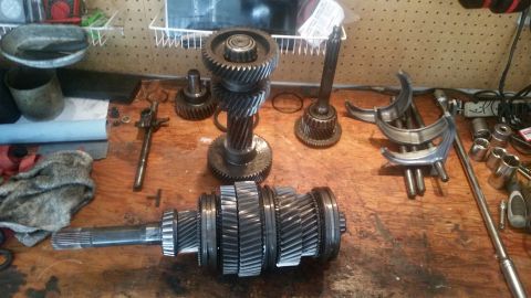 1985 Ford Bronco - A disassembled transmission lays on a wooden workbench. Several gears, shafts, and bearing cages are visible, along with tools and a rag.