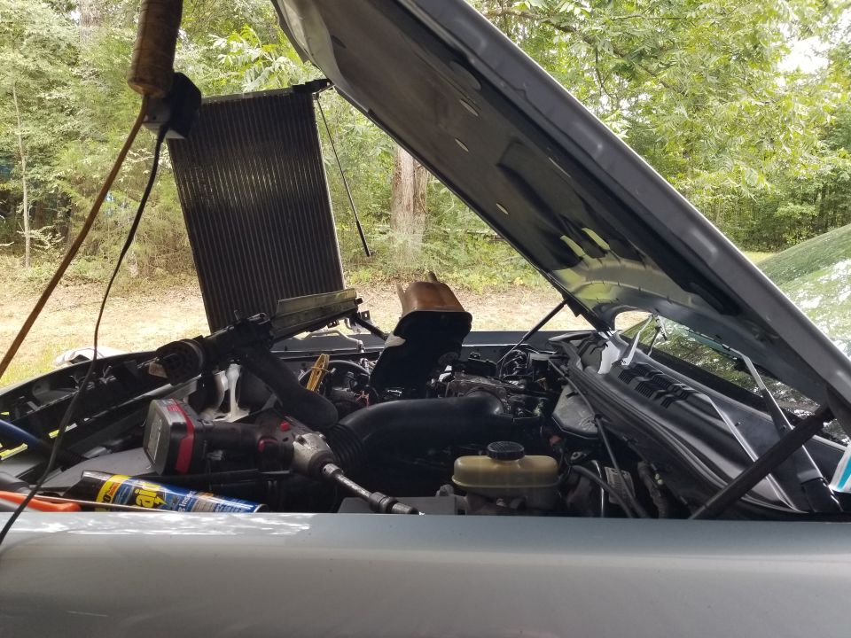 Silver sedan with hood up outdoors; radiator lifted out of engine bay, drill and tools resting on the engine.