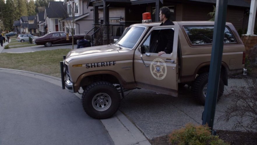 Tan Ford Bronco sheriff truck with red roof light and star door decal, man standing at open door on suburban street