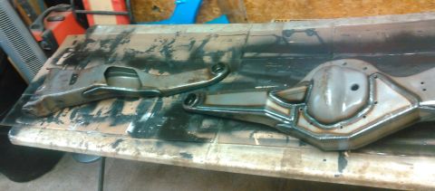 1992 Ford Bronco - A bare-metal radius arm and a stripped front axle housing with differential cover boss lie on a paint-splattered workbench in an OSB-walled…