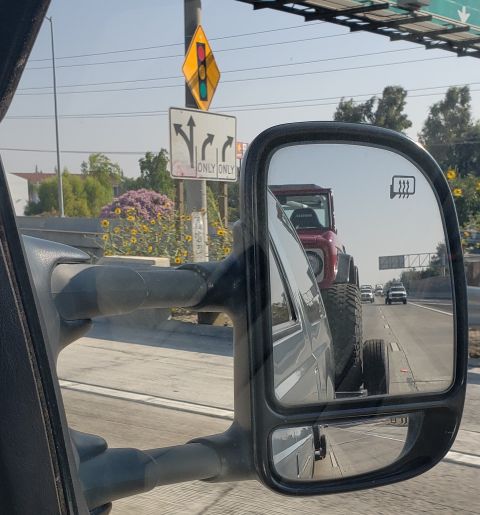 1968 Ford Bronco - A red Jeep Wrangler with large off-road tires is reflected in the side mirror of a dark-colored truck. The Jeep is being towed or is follow…