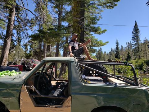 1968 Ford Bronco - A man sits on top of a heavily modified green off-road vehicle, holding a rope. The vehicle appears to be a custom build with a roll cage a…