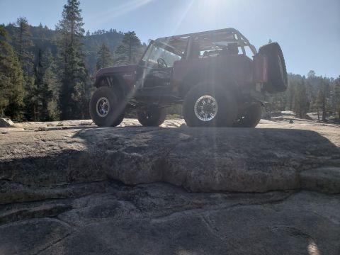 1968 Ford Bronco - A modified, dark red Ford Bronco with a roll cage and large off-road tires is parked on large rocks in a wooded, mountainous area. The spar…