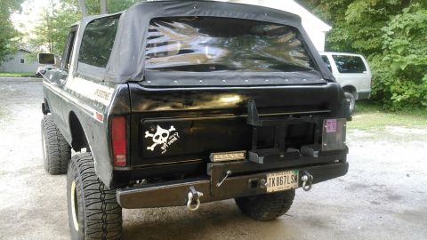1978 Ford Bronco - The rear of a black, lifted Ford Bronco with a soft top is shown parked outdoors on a gravel surface. Visible modifications include a custo…
