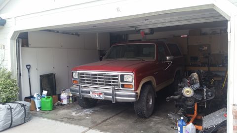 1985 Ford Bronco - A red 1980s Ford Bronco is parked inside a garage, partially obscuring a disassembled engine on a stand. Various items, including a green g…