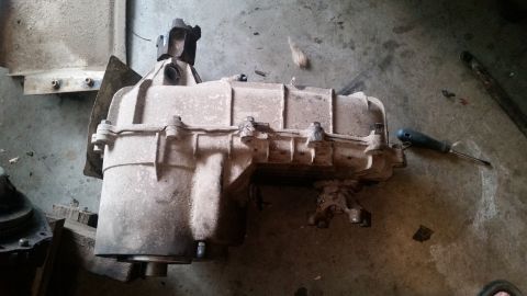 1985 Ford Bronco - A dusty transfer case is shown resting on a concrete floor, possibly in a garage or workshop. A screwdriver lies nearby.