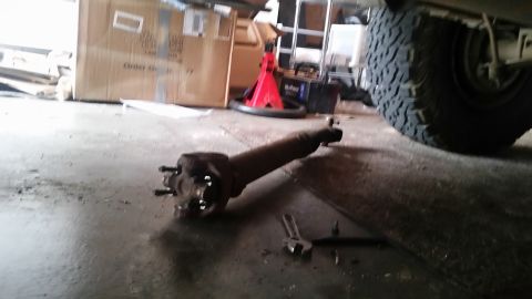1985 Ford Bronco - A detached automotive drive shaft with a U-joint is lying on a concrete floor in a garage. A large tire and part of the vehicle's frame are…