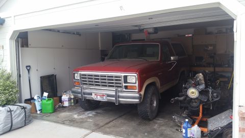 1985 Ford Bronco - A red 1980s Ford Bronco is parked inside a garage, partially obscured by a disassembled engine on a stand. Various tools, containers, and c…
