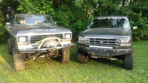 1992 Ford Bronco - Two lifted Ford Broncos parked front-to-camera on grass in front of dense trees, both carrying 'Broncos By Brandon' lettering. At left is a…