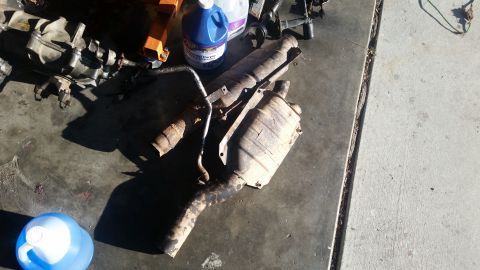 1985 Ford Bronco - Several exhaust system components, including catalytic converters and mufflers, are laid out on a concrete surface. A blue jug and a purple…