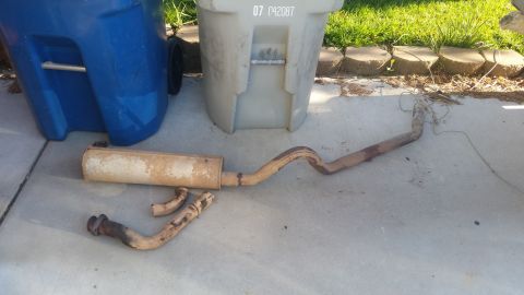 1985 Ford Bronco - A used exhaust system, including a muffler and pipes, lies on a concrete surface next to two large trash cans. The exhaust components are r…