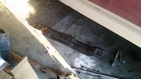 1985 Ford Bronco - A dark, rusty metal bracket lies on a concrete floor, illuminated by a strong shaft of sunlight. Tools, including a ratchet and socket, are…