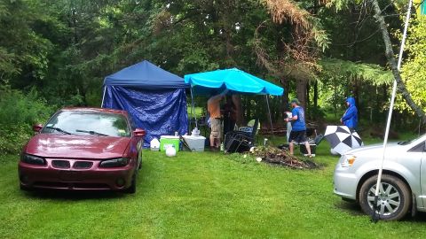 2000 Pontiac Grand Prix - A maroon Pontiac Grand Prix is parked on a grassy area in front of a wooded area with tents and people gathered under umbrellas.