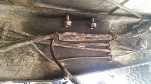 1985 Ford Bronco - A close-up view of rusty metal frame components with several bundled electrical wires and brake lines secured by clamps. Two bolts are visi…