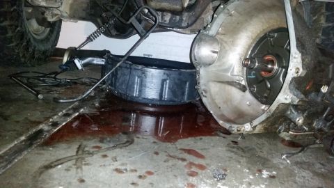 1985 Ford Bronco - Transmission fluid has spilled onto the garage floor, with a vehicle's undercarriage and a transmission component in view.