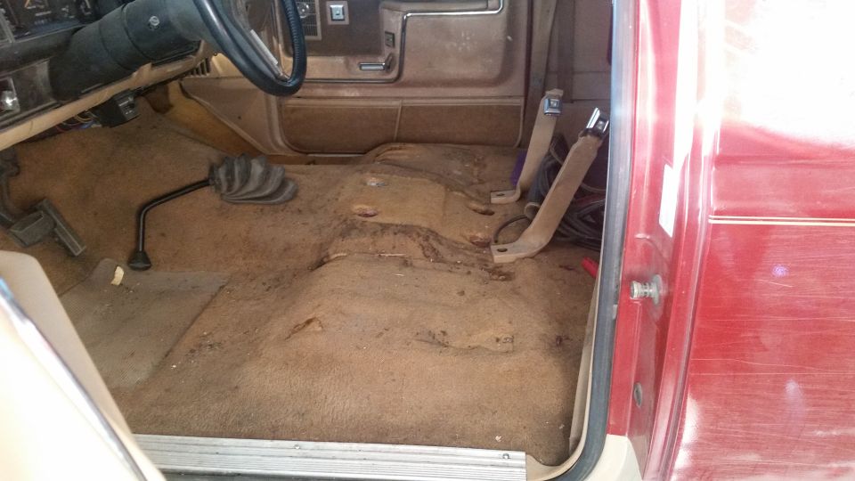 Interior of a red vehicle with worn brown carpet, exposed floor, and a manual shifter. Seat belts are visible