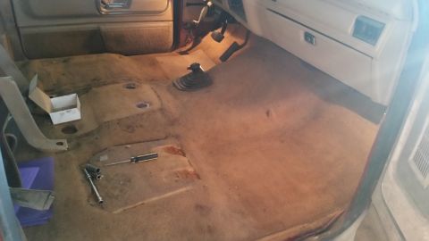 1985 Ford Bronco - The interior floor of a vehicle is shown with the carpet removed, revealing the transmission tunnel and some tools.