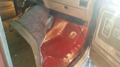 1985 Ford Bronco - The interior floor of a vehicle shows exposed red metal and a gray mat, with a circular plate on the floor.
