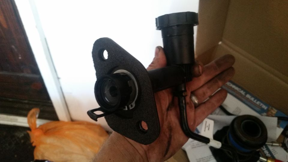 Hand holding a clutch master cylinder assembly with a black reservoir and mounting flange