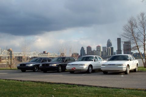 1998 Ford Crown Victoria - Four Ford Crown Victorias are parked in a lot with a city skyline visible in the distance.