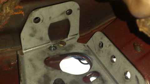 1985 Ford Bronco - Close-up view of a metal bracket with multiple holes and a red painted surface, likely part of a vehicle's undercarriage or engine bay.