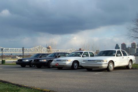 1998 Ford Crown Victoria - A line of four Ford Crown Victoria sedans are parked in a lot, with a bridge and city skyline visible.