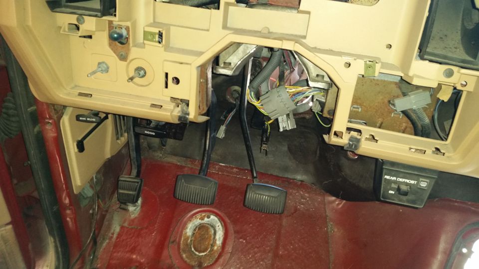 Vehicle interior showing dashboard, pedals, and wiring. Tan dashboard, red floor, and exposed wires are visible