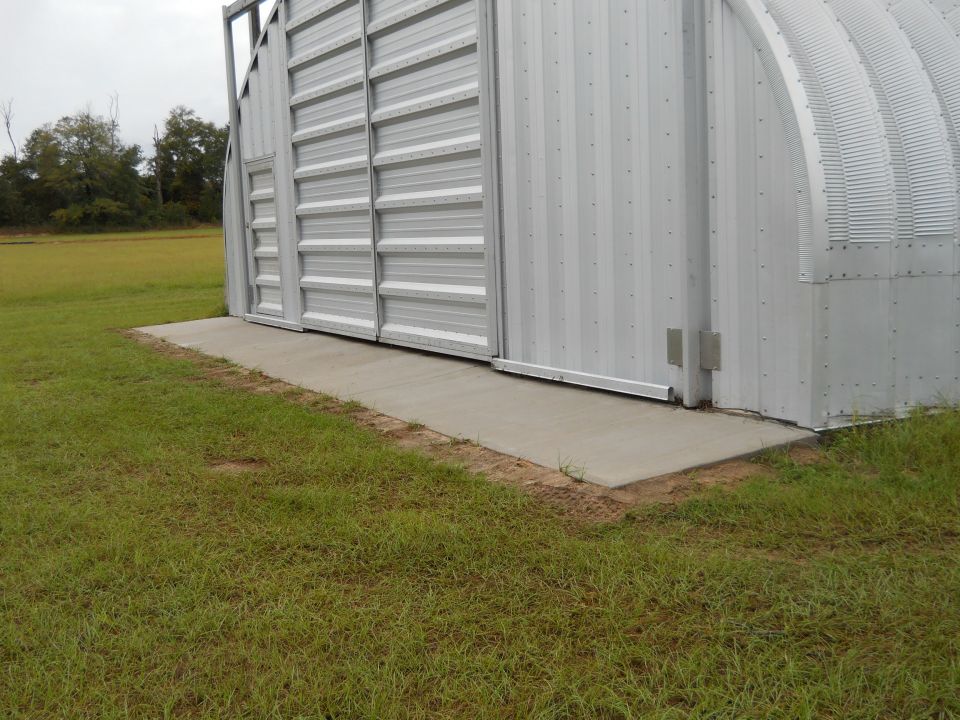 Side view of a large metal Quonset hut building with a concrete ramp leading to its sliding doors