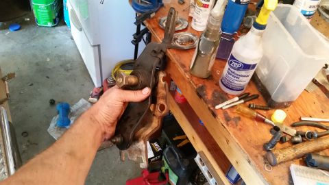 1985 Ford Bronco - A hand holds a transmission shifter linkage part over a workbench with tools and cleaning supplies.