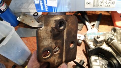 1985 Ford Bronco - A close-up view of a rusty metal bracket with a bolt and multiple holes, likely a vehicle part.