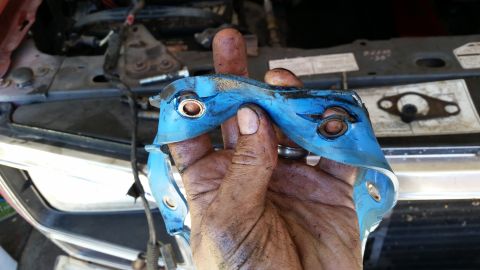1985 Ford Bronco - A dirty hand holds a blue gasket part in front of an engine bay, suggesting engine work.