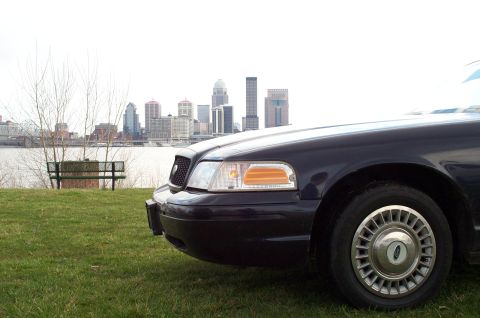 1998 Ford Crown Victoria - A dark 1998 Ford Crown Victoria is parked on a grassy hill overlooking a city skyline and river.