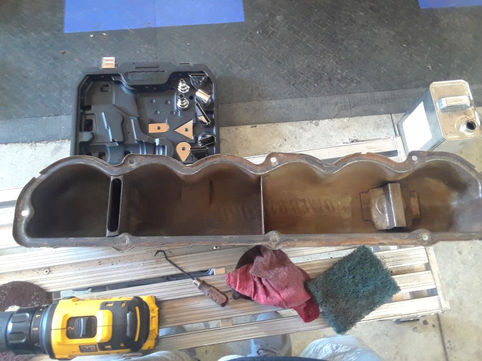 Engine valve cover with internal baffles, resting on a wooden pallet next to a drill and cleaning supplies
