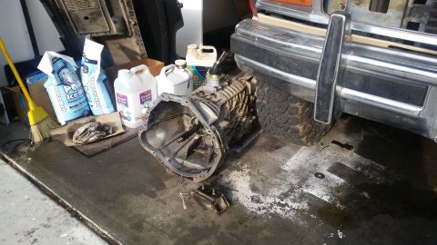 1985 Ford Bronco - A manual transmission is laid out on a garage floor next to bags of ice melt and containers of fluid, with the front bumper and tire of a v…