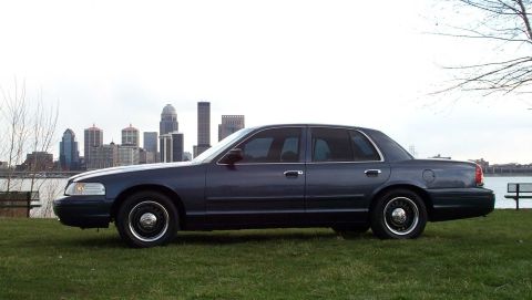 1998 Ford Crown Victoria - A 1998 Ford Crown Victoria sedan is shown from the side with a city skyline visible across the water.