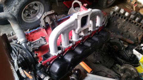 1985 Ford Bronco - A close-up view of a disassembled V8 engine, featuring a silver Offenhauser intake manifold bolted onto a red engine block with black valve…
