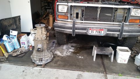 1985 Ford Bronco - A dirty, disassembled manual transmission is laid out on the ground next to the front end of a red 1980s Ford truck. Various containers of…