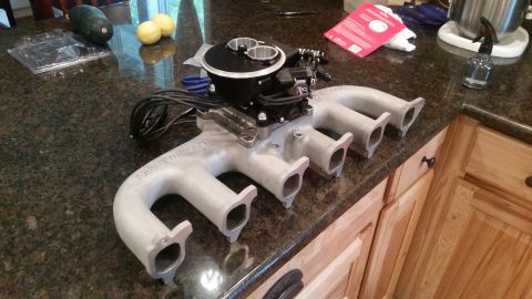 1985 Ford Bronco - A silver aluminum intake manifold with a black throttle body is displayed on a granite countertop. The manifold has four exhaust ports and…