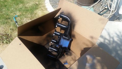 1985 Ford Bronco - A black exhaust manifold is placed inside a cardboard box, with some blue tape visible on the bottom of the box. The manifold has the text…