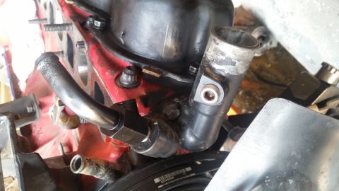 1985 Ford Bronco - A close-up view of a black thermostat housing and metal pipe attached to a red engine block. A serpentine belt is visible at the bottom of…