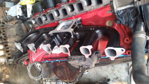 1985 Ford Bronco - A close-up view of a red V8 engine block with a black intake manifold installed. The exhaust manifolds are also black and are attached to t…