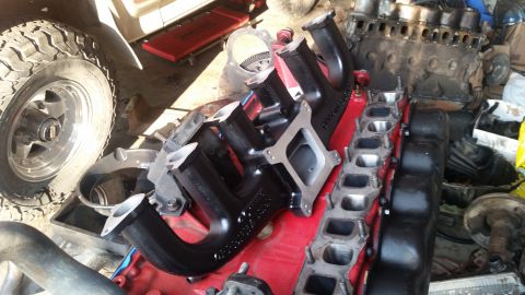 1985 Ford Bronco - A black aftermarket intake manifold is shown on top of a red engine block, with a large off-road tire and other engine parts in the backgro…