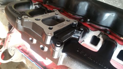 1985 Ford Bronco - A close-up view shows a black intake manifold with a silver adapter plate bolted to it. The intake manifold has visible text that appears t…
